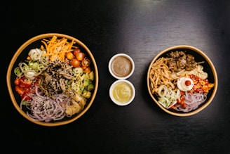 Two bowls filled with a variety of colorful ingredients are placed on a dark surface. The left bowl contains sliced vegetables such as carrots, cherry tomatoes, avocado cubes, sliced red onions, and mushrooms, garnished with sesame seeds. The right bowl also has a mix of vegetables, grilled meat, and a creamy sauce topping, accompanied by crunchy fried onions. Three small bowls of different sauces are arranged between the two larger bowls.