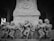 A detailed stone carving on an ancient pillar showcases a group of intricately sculpted figures in dynamic poses, each with distinct gestures and traditional attire. The scene captures a sense of historical and cultural richness.