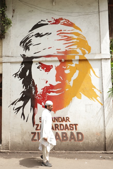 A mural painted on a wall features the face of a person with long hair and a headscarf, depicted in bold strokes of black, red, and orange. Below the mural, a man dressed in traditional white attire and a white cap is walking past, holding papers in one hand. The words 'Sikandar Zardast Zindabad' are inscribed beneath the mural.