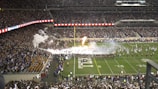 A large football stadium filled with spectators on both sides. A marching band is performing on the field, emitting white smoke and flames in a dramatic display. The crowd is actively engaged, capturing the excitement of a football game day.