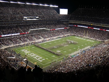 A large football stadium filled with spectators, featuring a marching band performing on the field. The stands are packed with people, and the stadium lights illuminate the area. The field displays the words 'TEXAS A&M'. The atmosphere is lively with a sense of anticipation.
