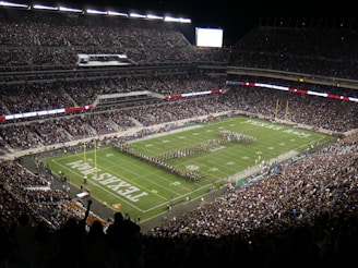 A large football stadium filled with spectators, featuring a marching band performing on the field. The stands are packed with people, and the stadium lights illuminate the area. The field displays the words 'TEXAS A&M'. The atmosphere is lively with a sense of anticipation.