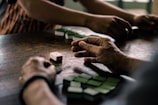 A group of hands engages in a game using domino tiles on a wooden table. The tiles are mostly green and are being arranged or played strategically by several players. The setting appears to be relaxed and informal, suggesting a casual gathering or a leisure activity.