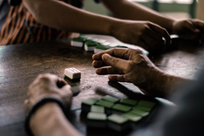 A group of hands engages in a game using domino tiles on a wooden table. The tiles are mostly green and are being arranged or played strategically by several players. The setting appears to be relaxed and informal, suggesting a casual gathering or a leisure activity.