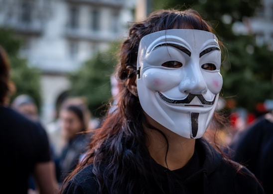 A person wearing a Guy Fawkes mask stands in a crowd. The mask is white with stylized features including a curled mustache and a goatee. The background is blurred with people and greenery, suggesting an outdoor setting.