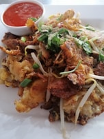 A plate of crispy fried seafood topped with green onions and cilantro. Bean sprouts are scattered throughout the dish, and a small bowl of red chili sauce is placed beside it.
