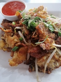 A plate of crispy fried seafood topped with green onions and cilantro. Bean sprouts are scattered throughout the dish, and a small bowl of red chili sauce is placed beside it.