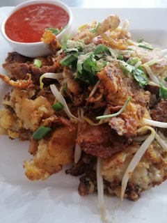 A plate of crispy fried seafood topped with green onions and cilantro. Bean sprouts are scattered throughout the dish, and a small bowl of red chili sauce is placed beside it.