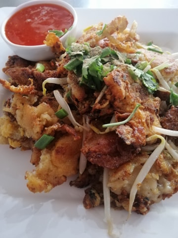 A plate of crispy fried seafood topped with green onions and cilantro. Bean sprouts are scattered throughout the dish, and a small bowl of red chili sauce is placed beside it.