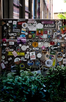 A street scene featuring a wall or surface covered with a large assortment of colorful stickers, each displaying various designs and texts. The scene is partially obscured by lush green foliage in the foreground. The stickers range from cartoonish graphics to text-based designs, contributing to an urban and artistic aesthetic.