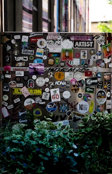 A street scene featuring a wall or surface covered with a large assortment of colorful stickers, each displaying various designs and texts. The scene is partially obscured by lush green foliage in the foreground. The stickers range from cartoonish graphics to text-based designs, contributing to an urban and artistic aesthetic.