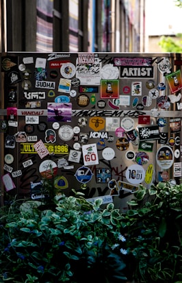 A street scene featuring a wall or surface covered with a large assortment of colorful stickers, each displaying various designs and texts. The scene is partially obscured by lush green foliage in the foreground. The stickers range from cartoonish graphics to text-based designs, contributing to an urban and artistic aesthetic.