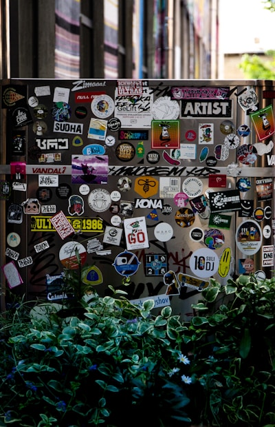 A street scene featuring a wall or surface covered with a large assortment of colorful stickers, each displaying various designs and texts. The scene is partially obscured by lush green foliage in the foreground. The stickers range from cartoonish graphics to text-based designs, contributing to an urban and artistic aesthetic.