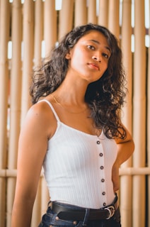 A person with curly hair is wearing a white sleeveless top with buttons and dark jeans, standing against a bamboo background.
