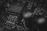 A close-up view of a circuit board featuring a microchip with visible alphanumeric inscriptions. The intricate details of the board include soldered components and electronic pathways, highlighting its complex design.