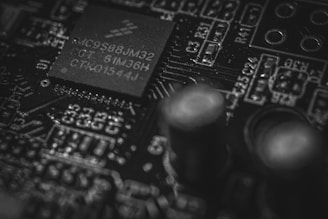 A close-up view of a circuit board featuring a microchip with visible alphanumeric inscriptions. The intricate details of the board include soldered components and electronic pathways, highlighting its complex design.