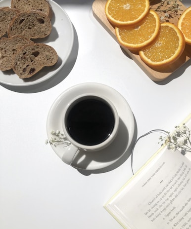A neatly arranged breakfast setup on a white surface, featuring a plate with slices of brown bread, a wooden board with sliced oranges, a cup of black coffee, and an open book with small white flowers as decoration.