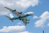 A large commercial airplane with distinctive turquoise and white livery is flying in a clear blue sky with scattered white clouds. The plane's engines and wings are prominently visible, and the aircraft is angled slightly upwards as if ascending.