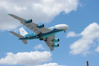 A large commercial airplane with distinctive turquoise and white livery is flying in a clear blue sky with scattered white clouds. The plane's engines and wings are prominently visible, and the aircraft is angled slightly upwards as if ascending.