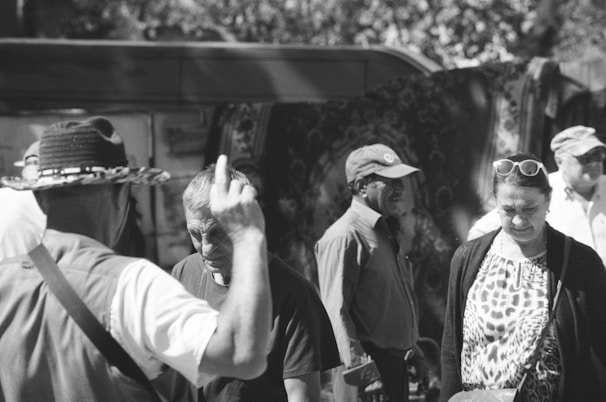 A black and white image features a group of people in a casual outdoor setting. The individuals are dressed in varied clothing, including hats and sunglasses, suggesting a warm, sunny day. They appear to be engaged in conversation or personal contemplation, with some facing each other while others look down or away.