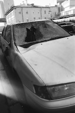 A damaged car with a shattered windshield parked on the side of a street. The surrounding buildings are residential or commercial structures with multiple floors.