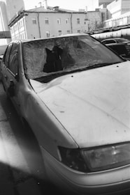 A damaged car with a shattered windshield parked on the side of a street. The surrounding buildings are residential or commercial structures with multiple floors.