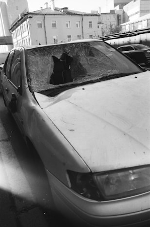 A damaged car with a shattered windshield parked on the side of a street. The surrounding buildings are residential or commercial structures with multiple floors.