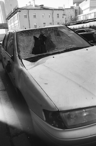 A damaged car with a shattered windshield parked on the side of a street. The surrounding buildings are residential or commercial structures with multiple floors.