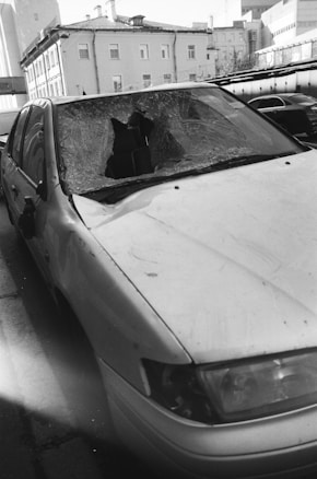 A damaged car with a shattered windshield parked on the side of a street. The surrounding buildings are residential or commercial structures with multiple floors.