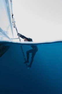 A person clad in a wetsuit is partially submerged in deep blue water, climbing a ladder attached to a boat. The waterline bisects the image, creating a stark contrast between the marine environment below and the bright sky above.