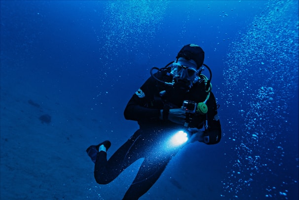 A scuba diver is underwater, surrounded by a deep blue sea. They are equipped with diving gear including a mask, breathing apparatus, and wetsuit. The diver is holding a flashlight, illuminating the water around them as bubbles rise to the surface.