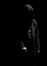 A mysterious figure is cloaked in a dark robe with a hood, obscuring their face in deep shadows. A large wooden cross hangs around their neck. The dim lighting creates stark contrasts, highlighting only parts of the figure's form, particularly the hands and the cross.