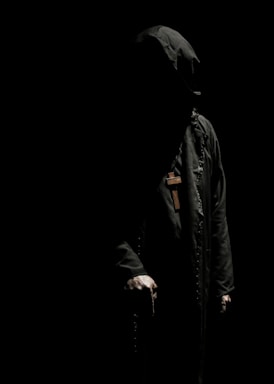 A mysterious figure is cloaked in a dark robe with a hood, obscuring their face in deep shadows. A large wooden cross hangs around their neck. The dim lighting creates stark contrasts, highlighting only parts of the figure's form, particularly the hands and the cross.