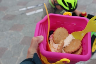 A pink toy basket filled with cookies and a plastic yellow spork, held by a hand. A child wearing a neon green helmet is in the background with a blurry view.