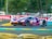A racing car with number 85 and vibrant purple and orange colors speeds along a track. The car features distinctive sponsorship logos and decals. The background includes blurred advertisements and greenery, indicating movement.