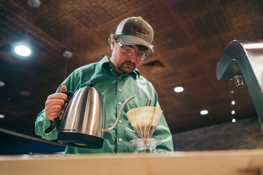 A person wearing a green shirt and cap is carefully pouring hot water from a stainless steel kettle into a coffee dripper placed over a glass carafe. The setting appears to be indoors with a warm, dimly lit ambiance, suggested by the ceiling lights.