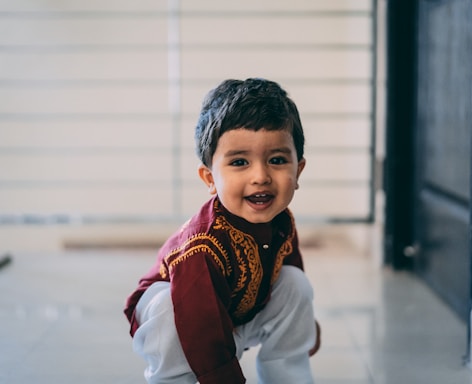 A young child wearing a traditional outfit with intricate embroidery, smiling and crouching indoors. The background is softly blurred, highlighting the child as the central focus.