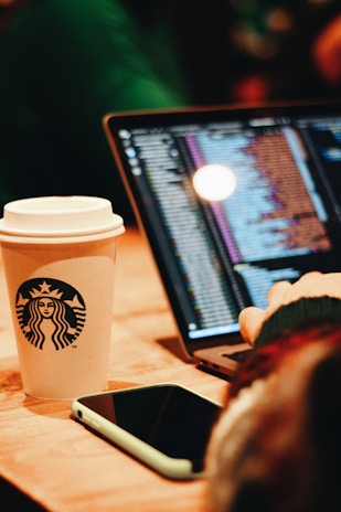 A close-up of a person using a laptop, with programming code displayed on the screen. A smartphone lies on the wooden table next to a Starbucks coffee cup with a logo. The atmosphere appears warm with soft lighting.