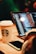 A close-up of a person using a laptop, with programming code displayed on the screen. A smartphone lies on the wooden table next to a Starbucks coffee cup with a logo. The atmosphere appears warm with soft lighting.