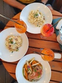 A wooden table set with three plates of pasta and meat dishes accompanied by vibrant drinks. Fresh ingredients and garnishes are artfully arranged on each plate, and the drinks appear to be colorful cocktails. The setting suggests an outdoor dining experience.