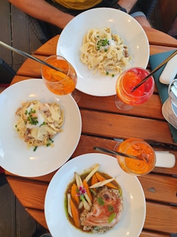 A wooden table set with three plates of pasta and meat dishes accompanied by vibrant drinks. Fresh ingredients and garnishes are artfully arranged on each plate, and the drinks appear to be colorful cocktails. The setting suggests an outdoor dining experience.