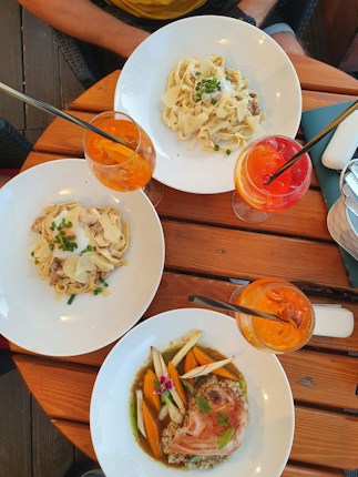 A wooden table set with three plates of pasta and meat dishes accompanied by vibrant drinks. Fresh ingredients and garnishes are artfully arranged on each plate, and the drinks appear to be colorful cocktails. The setting suggests an outdoor dining experience.