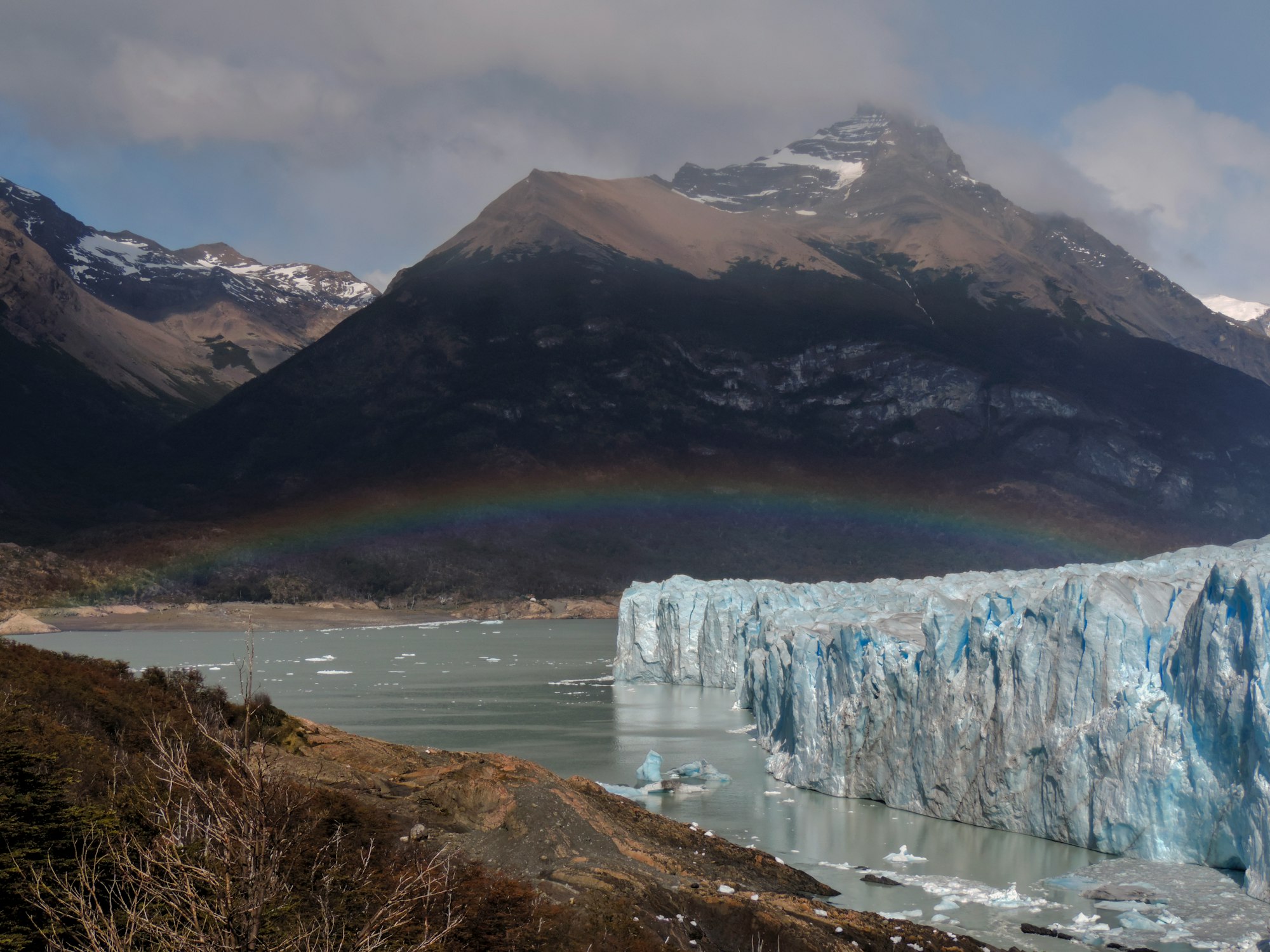 A rainbow over a lake with a glacier, tall snowy mountains are in the background with wisps of cloud.