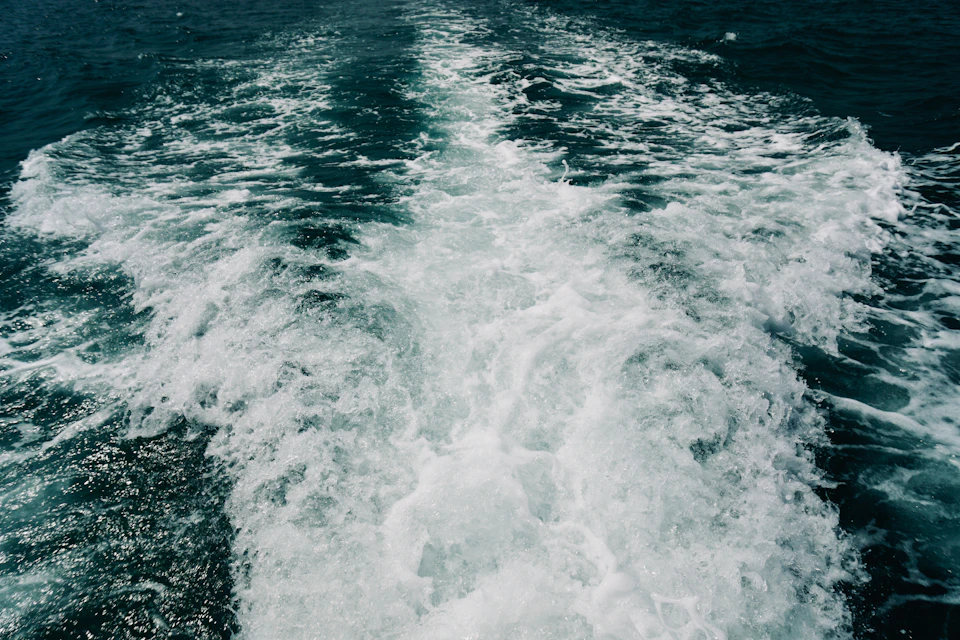 The image depicts a large body of water with a wake trailing behind a boat, creating a trail of white foam that contrasts against the deep blue color of the water. The boat is positioned on the left side of the frame and appears to be moving towards the right, leaving behind a distinct wake as it cuts through the water.
