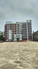 A multi-story building under construction with red and white exterior colors. The foreground is an open, sandy area, possibly indicating ongoing construction work. Scaffolding and construction materials are visible, along with a few figures, likely workers, near the building.