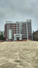 A multi-story building under construction with red and white exterior colors. The foreground is an open, sandy area, possibly indicating ongoing construction work. Scaffolding and construction materials are visible, along with a few figures, likely workers, near the building.