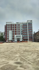 A multi-story building under construction with red and white exterior colors. The foreground is an open, sandy area, possibly indicating ongoing construction work. Scaffolding and construction materials are visible, along with a few figures, likely workers, near the building.