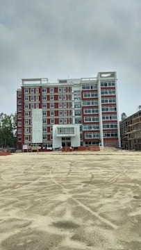 A multi-story building under construction with red and white exterior colors. The foreground is an open, sandy area, possibly indicating ongoing construction work. Scaffolding and construction materials are visible, along with a few figures, likely workers, near the building.