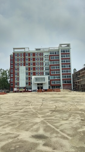 A multi-story building under construction with red and white exterior colors. The foreground is an open, sandy area, possibly indicating ongoing construction work. Scaffolding and construction materials are visible, along with a few figures, likely workers, near the building.
