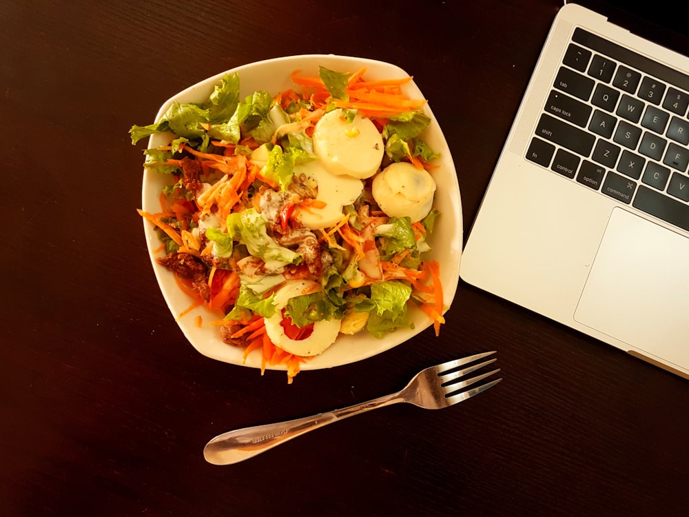 회색 노트북 컴퓨터 옆에 야채 샐러드가 있는 흰색 세라믹 그릇